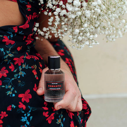 Person holding a bottle of perfume labeled 'Romantic' with white flowers in the background