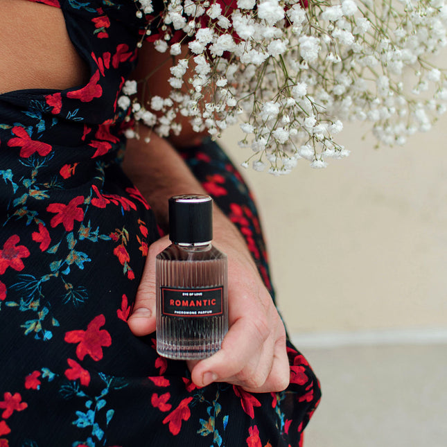 Person holding a bottle of perfume labeled 'Romantic' with white flowers in the background