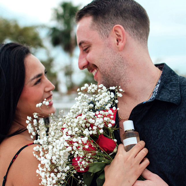 Man holding a bouquet of flowers and a after dark pheromone bottle, standing close to a woman outdoors.