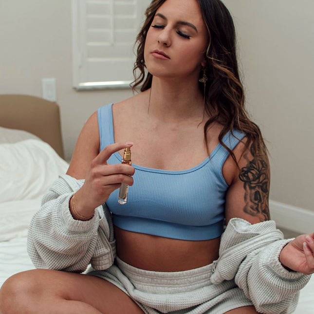 Woman in a blue sports bra holding a pheromone perfume bottle indoors.